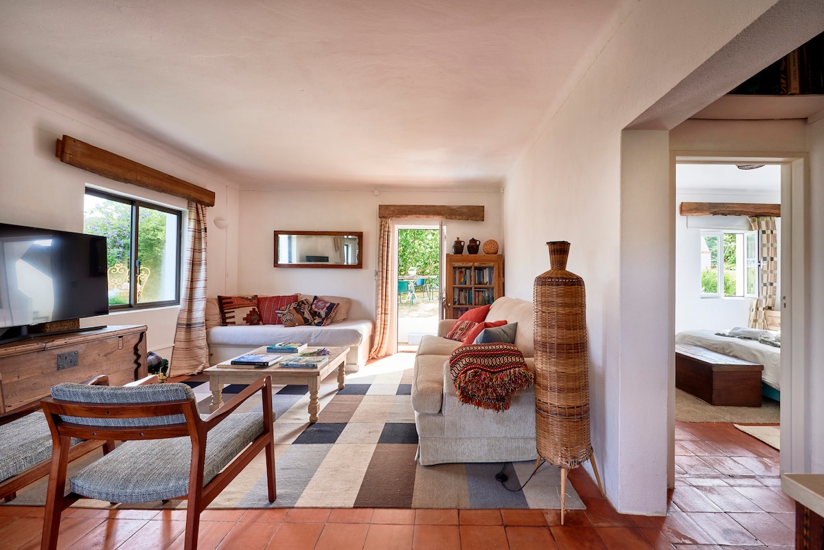 A spacious living area features large windows that invite natural light, framed by soft curtains. A comfortable sofa and chairs provide seating, with a coffee table at the center. A rug adds warmth to the terracotta-tiled floor, while a view of greenery is visible through the open doorway.