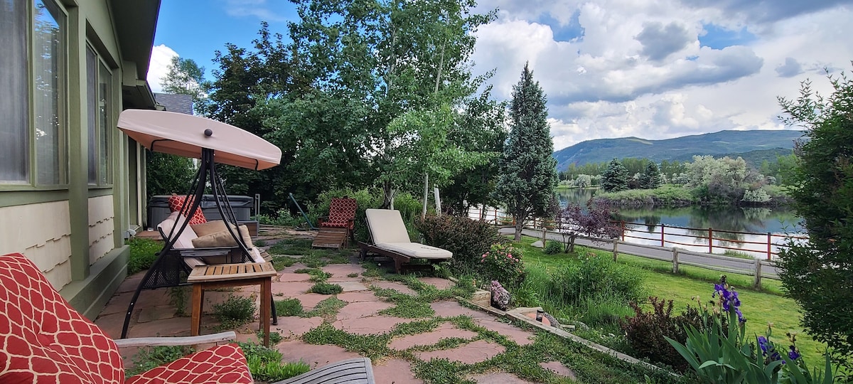 A peaceful outdoor patio features lounge chairs and a shaded reclining sunbed on a stone pathway, surrounded by landscaped greenery. The view includes a serene lake and mountains in the background, providing a tranquil setting for relaxation.