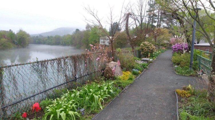 A peaceful pathway lined with vibrant greenery and blooming flowers leads along the water's edge. The serene lake is visible, surrounded by trees and soft morning mist, creating a calm and natural setting for leisurely strolls.
