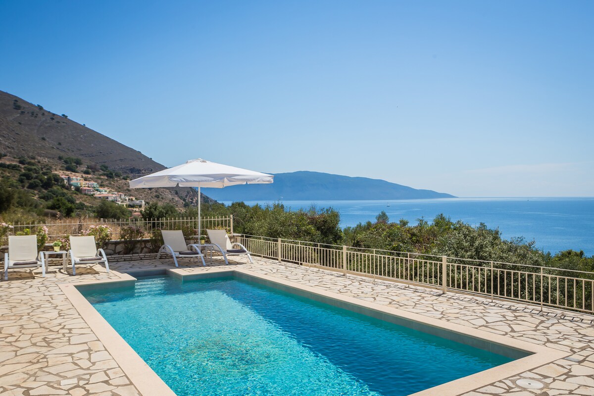 A serene swimming pool features clear blue water, set against a backdrop of rolling hills and an expansive sea view. Sun loungers with umbrellas are positioned around the pool area, inviting relaxation while enjoying the natural surroundings.