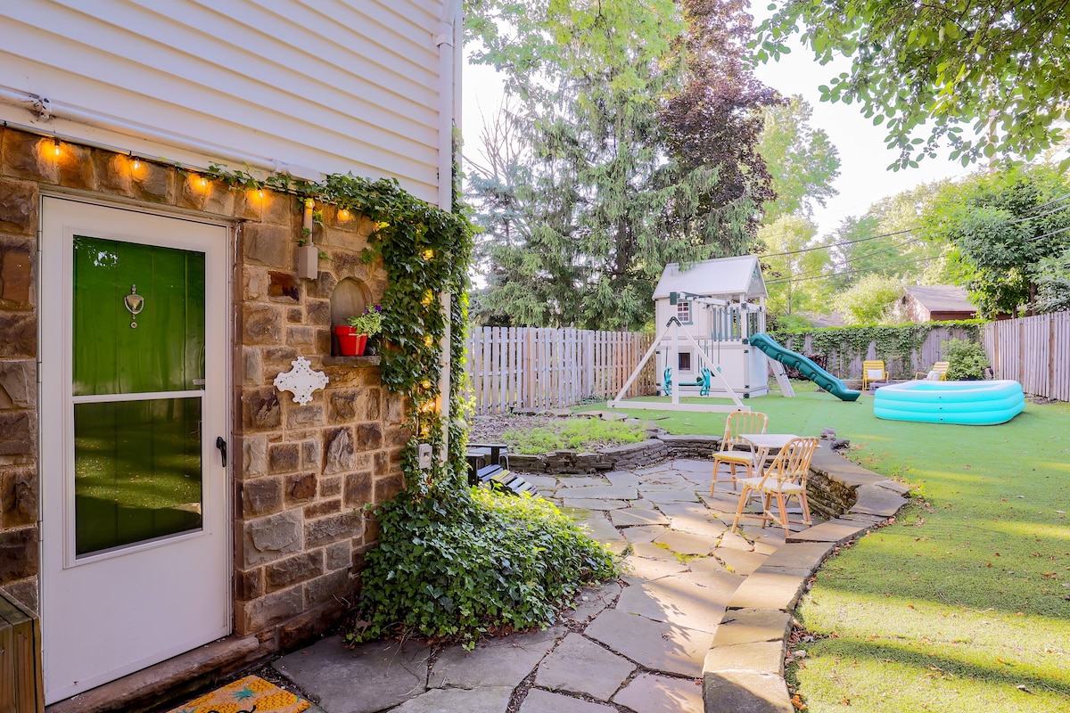 A stone pathway leads to a welcoming entrance, surrounded by greenery. The yard features a playset with a slide, seating areas, and a small inflatable pool. Lush grass fills the space, creating an inviting atmosphere for outdoor activities.