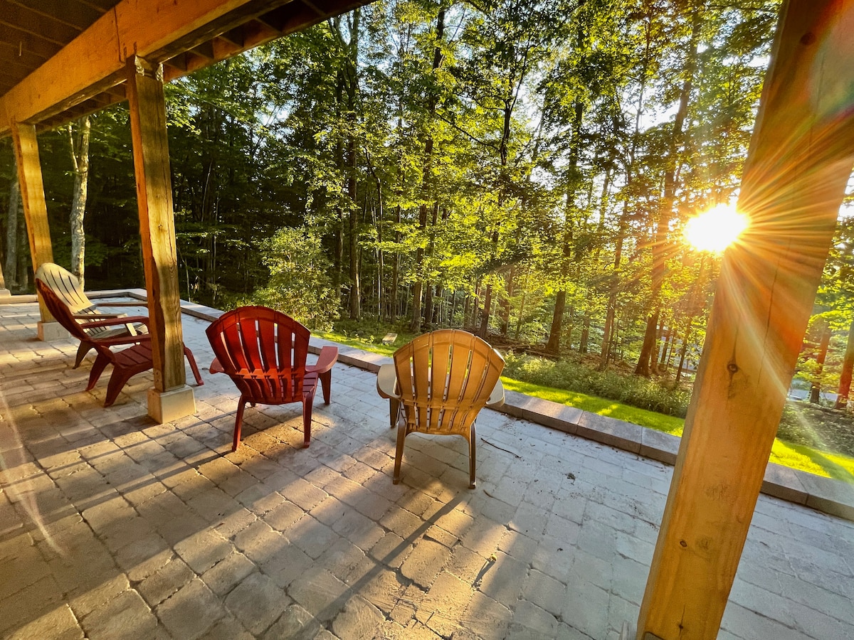 A spacious patio area features several Adirondack chairs arranged for relaxation. Sunlight filters through the trees, casting warm rays onto the stone surface and creating a serene outdoor setting. The lush greenery provides a tranquil backdrop.