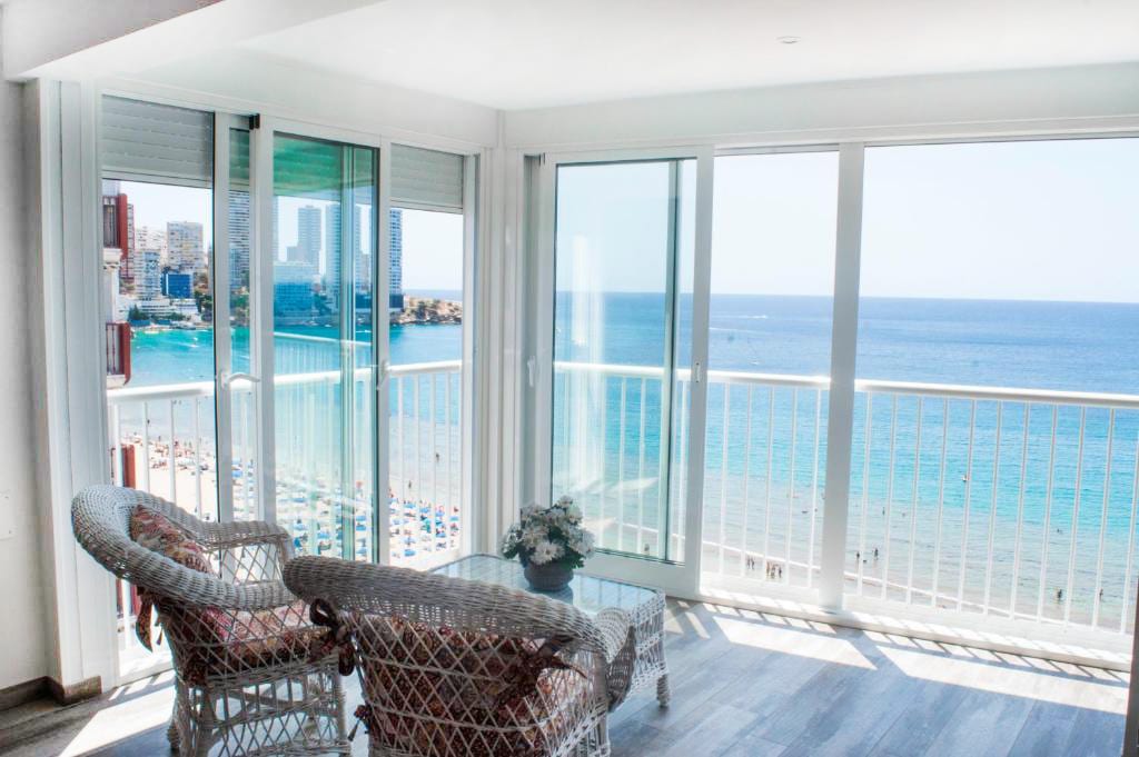 A bright living area features large glass doors that open to a stunning view of the sea. Two woven chairs and a glass table offer a comfortable spot to relax while the vibrant coastline can be seen through the expansive windows.