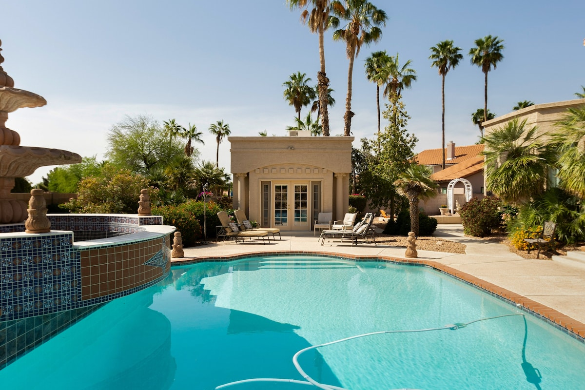 A private casita with French doors is visible beyond a serene pool. The pool area features tiled edges and is surrounded by tropical landscaping including palm trees and various plants, creating a relaxed outdoor setting.