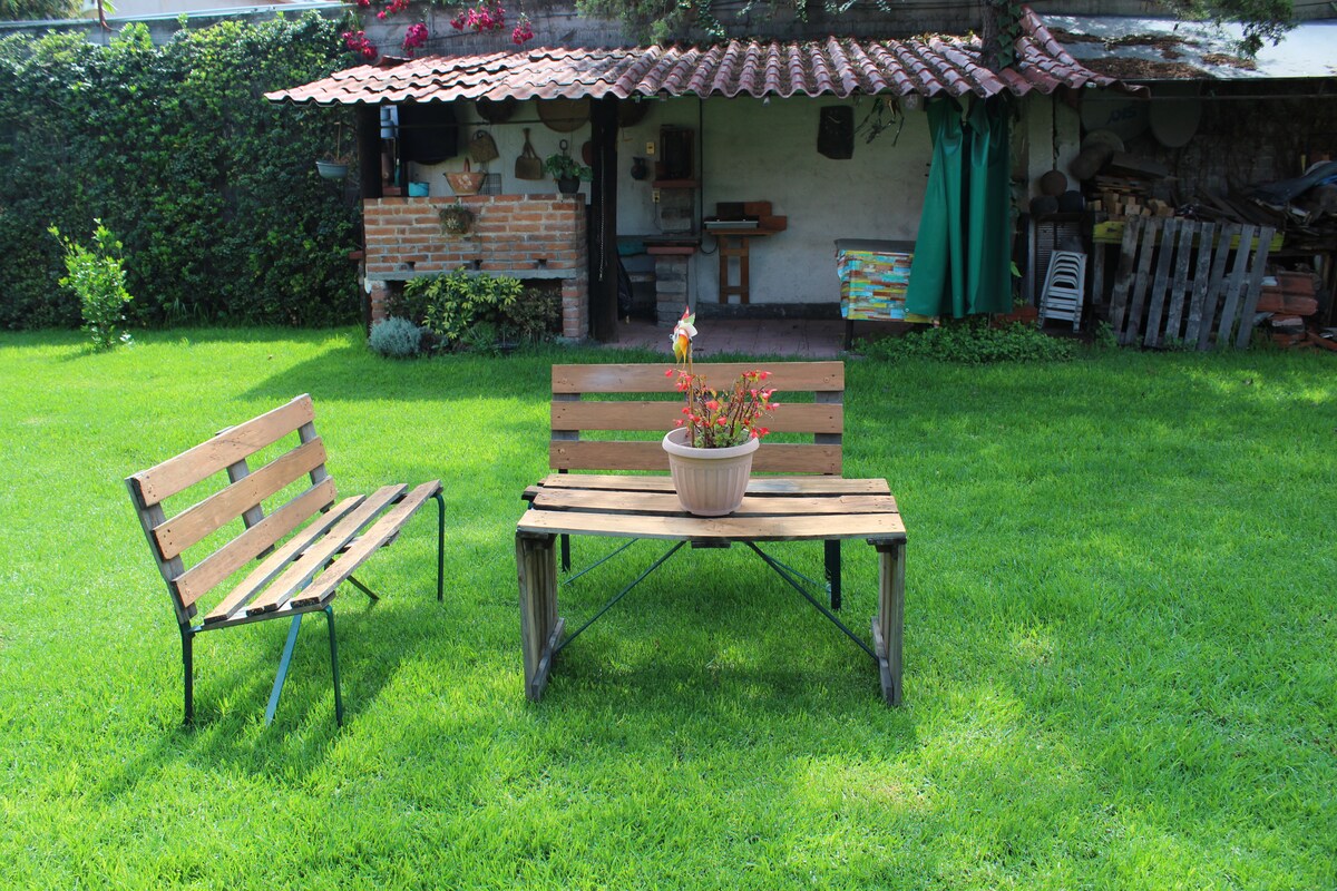 An inviting outdoor seating area features a wooden table accompanied by two benches. A potted plant sits at the table's center. Lush green grass surrounds the seating, with a background displaying a rustic outdoor kitchen and a variety of gardening tools, enhancing the tranquil garden setting.