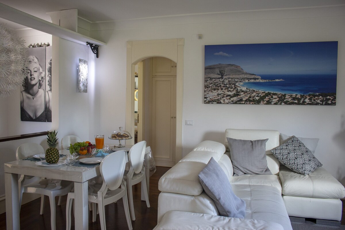 A bright living area is present, featuring a comfortable white sectional sofa adorned with a decorative pillow. A dining table is set for four, with a fruit centerpiece. A large artwork depicting a scenic coastal view is displayed on the wall.
