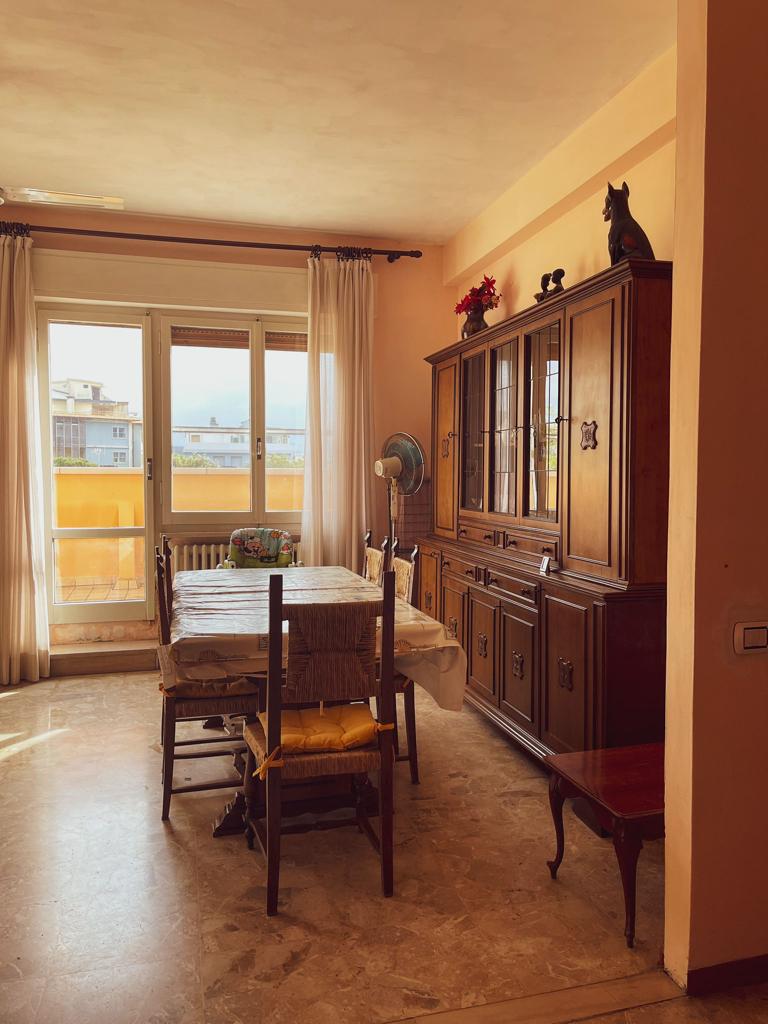 A dining area is shown with a wooden table surrounded by five chairs, one featuring a yellow cushion. A wooden cabinet stands against the wall, displaying decorative items. Natural light enters through large glass doors leading to a balcony, highlighting the room's spaciousness.