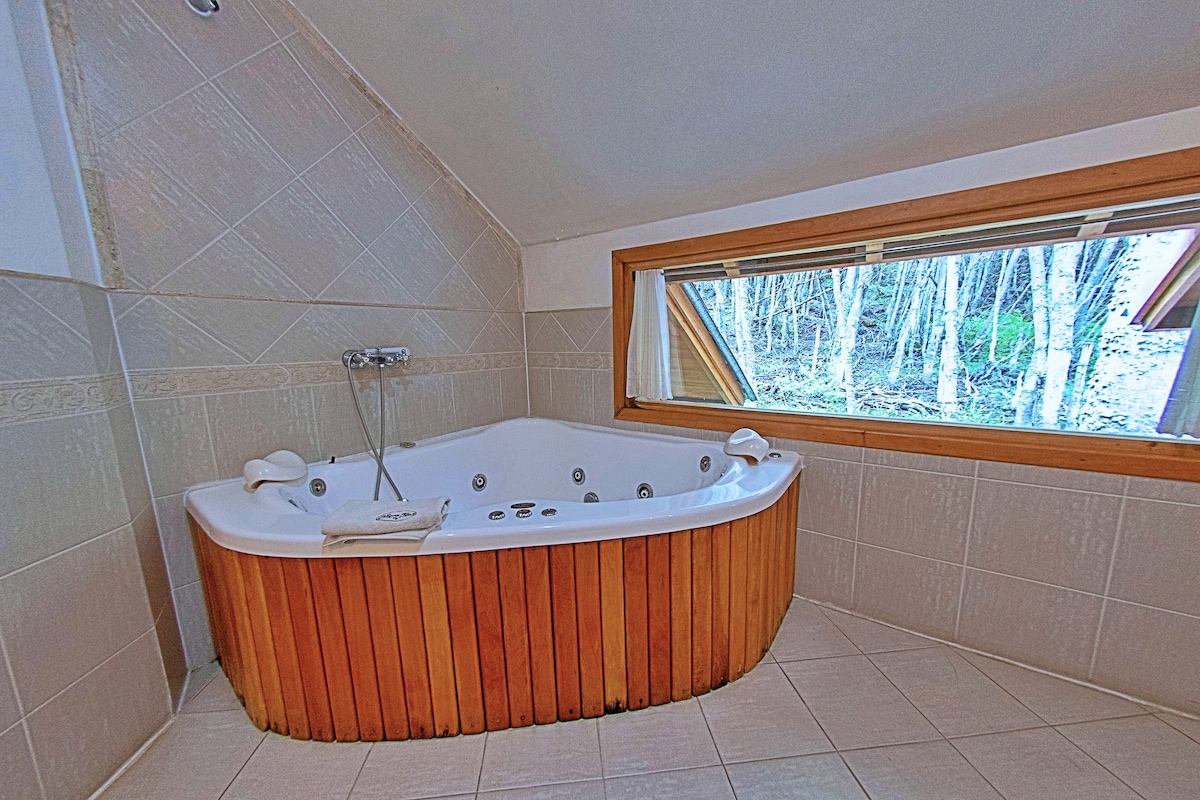 A spacious jacuzzi is positioned beside a large window, offering views of the surrounding forest. Wooden paneling encases the jacuzzi, which features multiple jets and controls. Light-colored tiles extend across the floor and walls, creating a serene atmosphere.