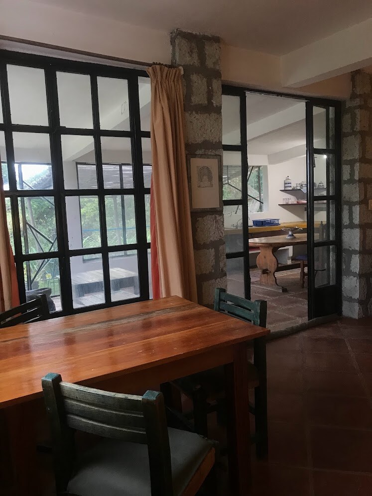 A wooden dining table with chairs is placed within a bright and airy space. Large glass doors provide expansive views of the outdoors, while natural light illuminates the interior. An adjacent room features a workspace with shelves, contributing to the open and inviting atmosphere.