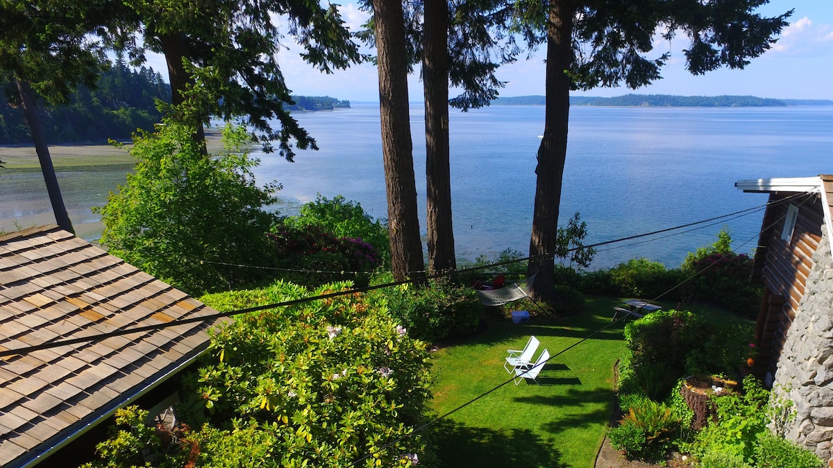 A serene view of Puget Sound emerges from a lush green garden, flanked by tall trees. A charming outdoor space is visible, featuring a single white lounge chair positioned on the manicured lawn, with tranquil water and distant mountains in the background.