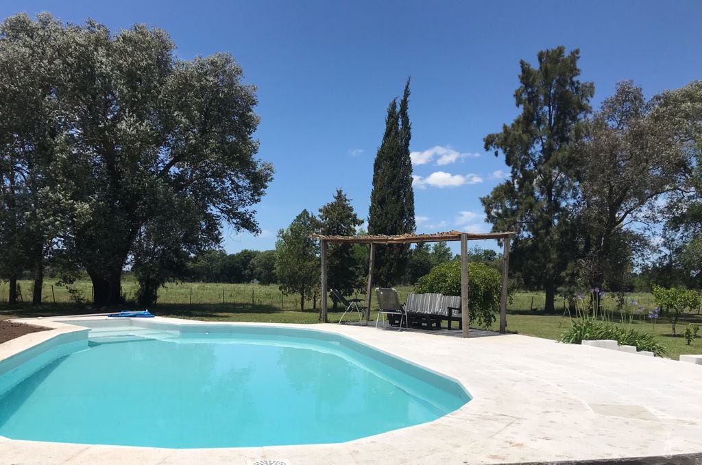 A clear blue swimming pool is surrounded by a stone deck, with a wooden pergola and seating area visible nearby. Lush greenery and trees create a natural backdrop, emphasizing the tranquil outdoor setting under a bright blue sky.