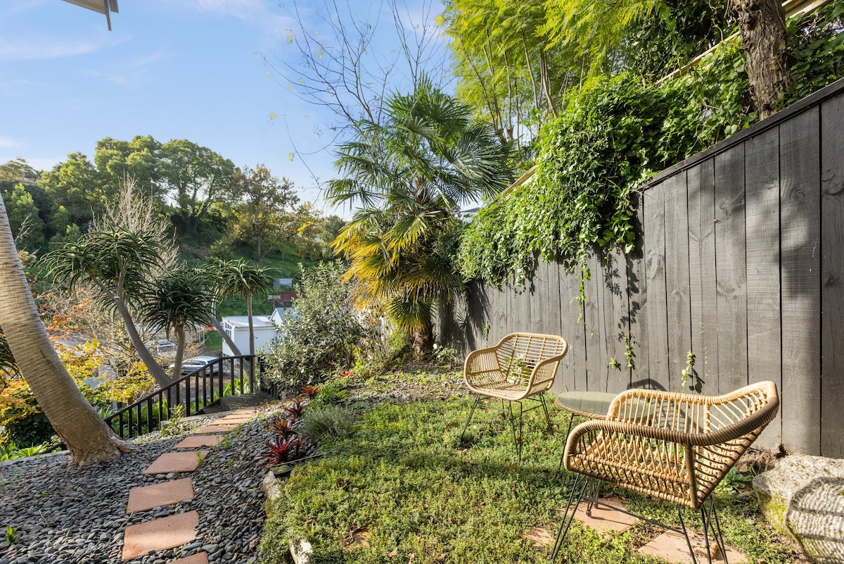 A private garden area features two rattan chairs positioned on a grassy space, surrounded by a variety of plants and shrubs. The wall behind is lined with greenery, providing a serene outdoor retreat. A pathway made of stone pavers leads through the area, enhancing accessibility.