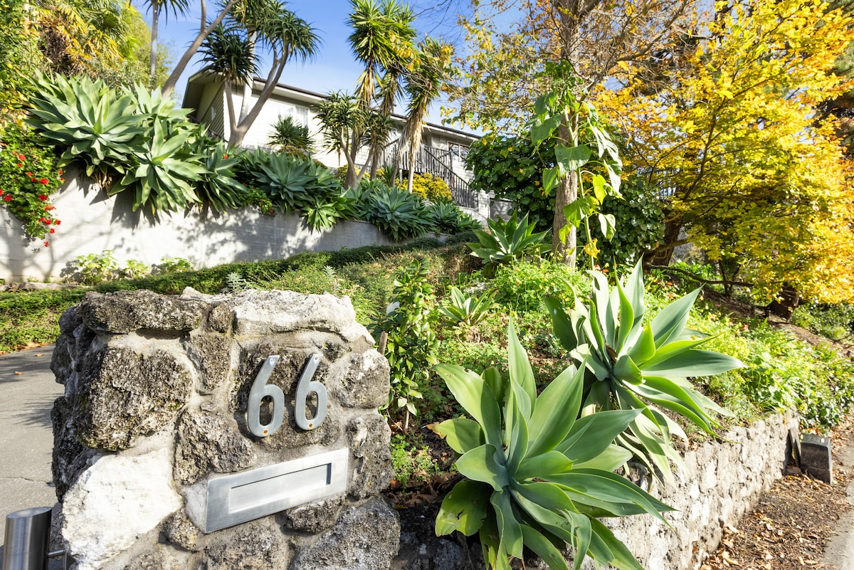 A stone wall with a modern mailbox features the number 66 prominently. Lush greenery including succulents and tropical plants surrounds the entrance, creating a vibrant scene. The backdrop showcases a charming single-story building nestled among foliage, illuminated by warm sunlight.