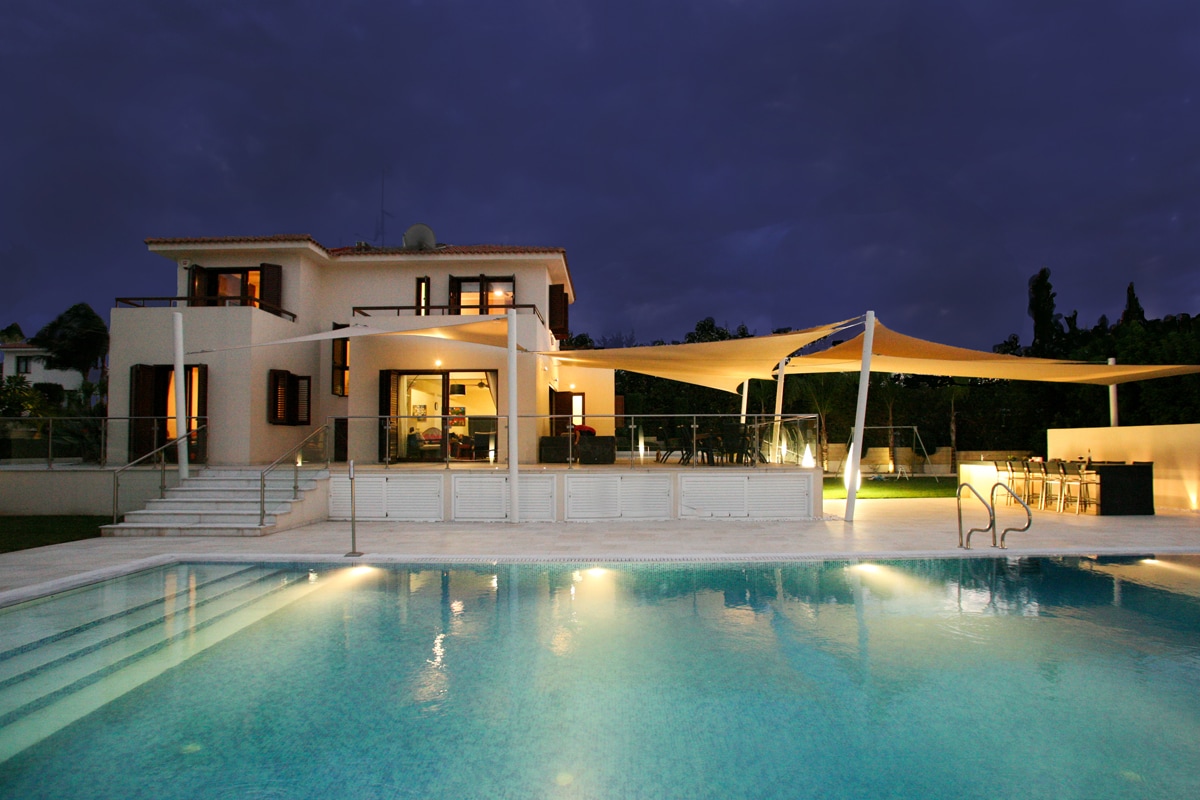 The exterior of the villa is presented at twilight, showcasing a modern structure with multiple balconies and large windows. The inviting overflow pool reflects the soft lighting from the terrace, while architectural sails provide shade over the outdoor dining area.