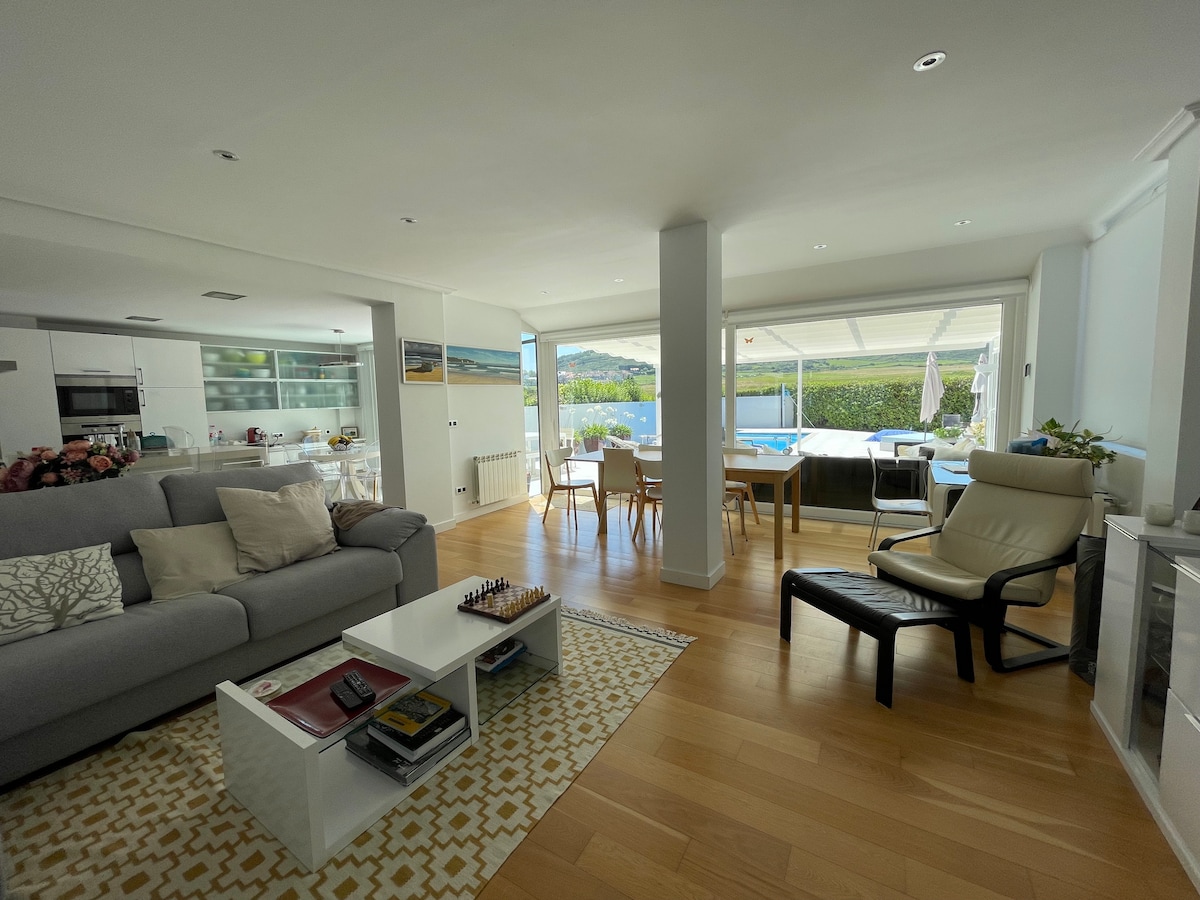 A spacious living area features a light-colored sofa and a low coffee table with games. Nearby, a dining space with wooden chairs is seen next to large windows that overlook the private pool area and greenery, creating an open, bright environment.