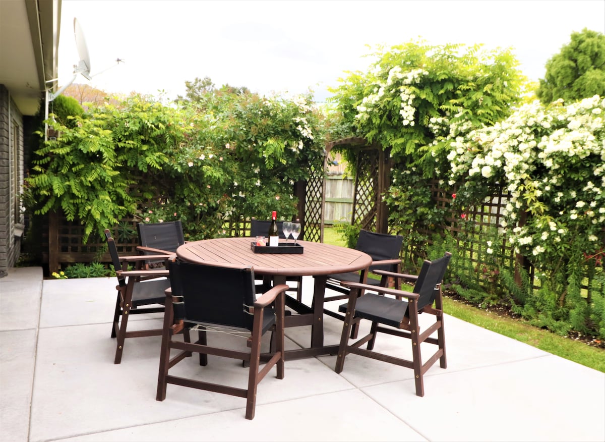 An outdoor dining area is displayed, featuring a round wooden table surrounded by four comfortable chairs. Lush greenery and flowering vines provide a natural backdrop, while a bottle of wine and two glasses sit elegantly on the table, inviting relaxation and conversation.