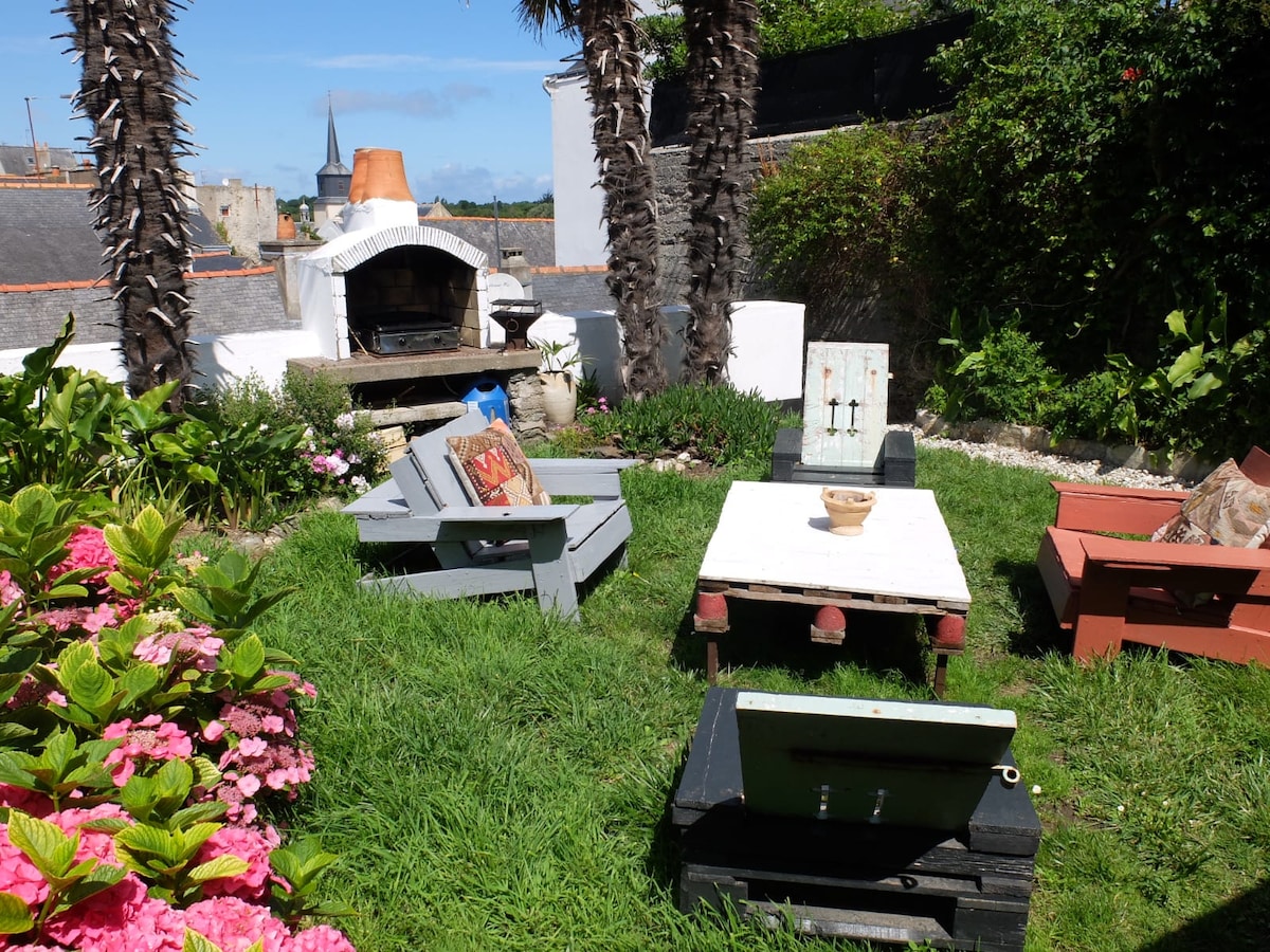 A charming garden area features several outdoor seating arrangements, including wooden chairs and a central table. Lush green grass and blooming flowers surround the space. A traditional fireplace stands in the background, providing an inviting focal point for outdoor gatherings.