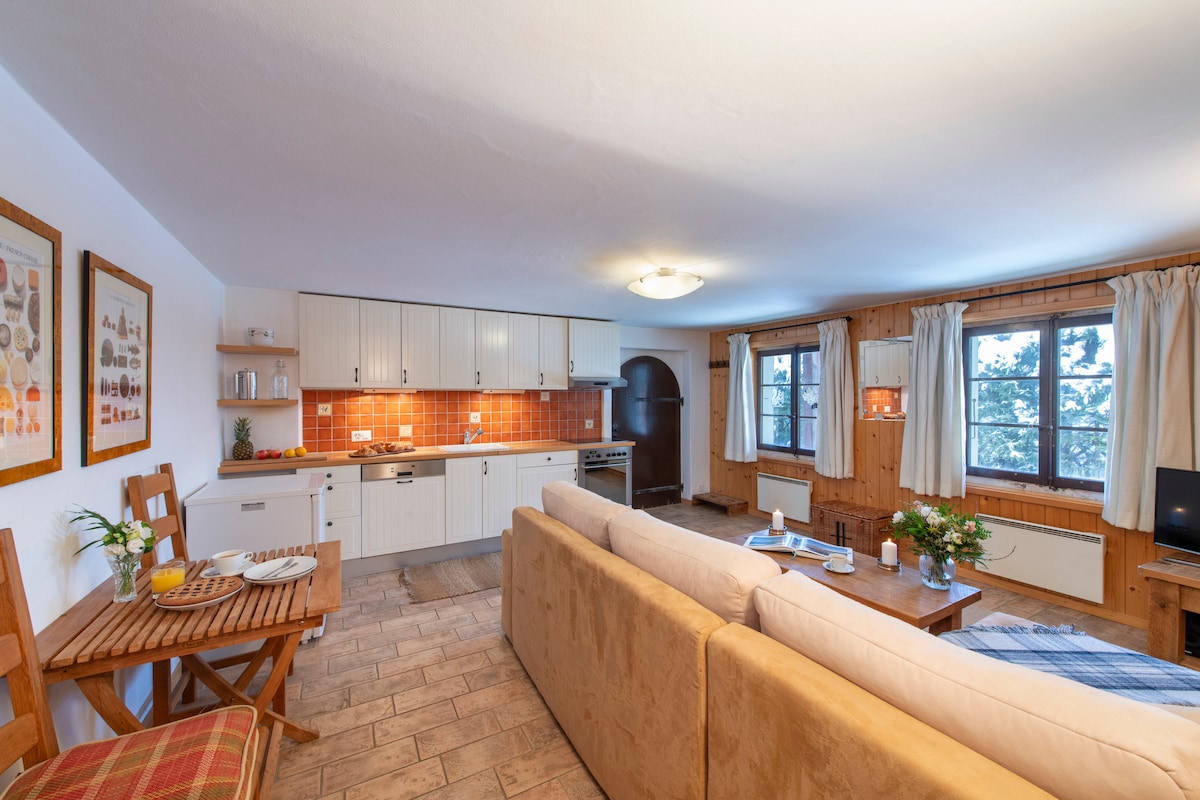 An open plan living area features a spacious beige sofa, complemented by wooden furniture. The kitchen, equipped with modern appliances and ample storage, is accentuated by orange tiled backsplash. Natural light fills the room through large windows, with light curtains enhancing the inviting ambiance.