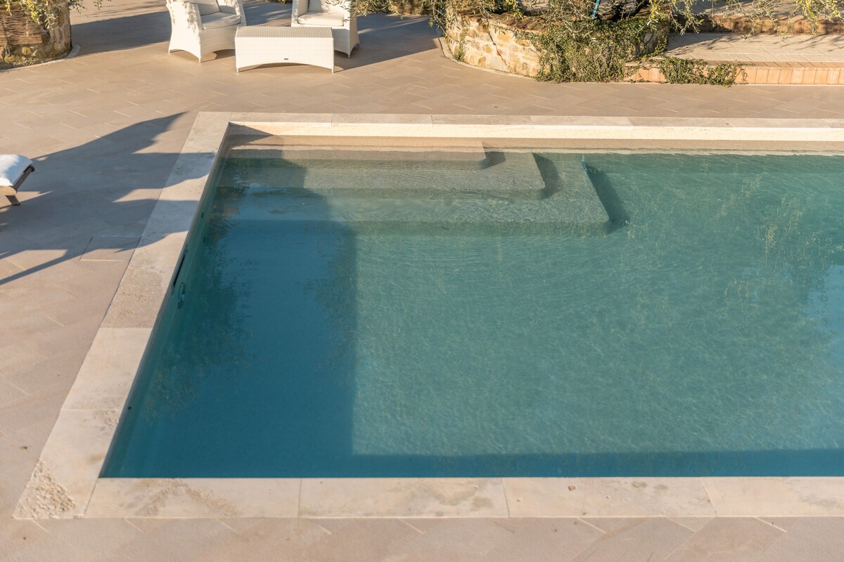 The inviting swimming pool features clear, blue water, with sunlight creating reflective patterns on the surface. Surrounding the pool are beige stone tiles, and the pool's edge is lined with a smooth light-colored coping, enhancing the overall tranquil setting.