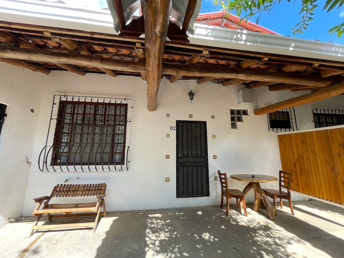 An outdoor seating area is presented, featuring a wooden bench and a round table with two chairs. The walls are painted white, complemented by wooden beams overhead. A black door with decorative hardware is visible, contributing to the space's rustic charm.