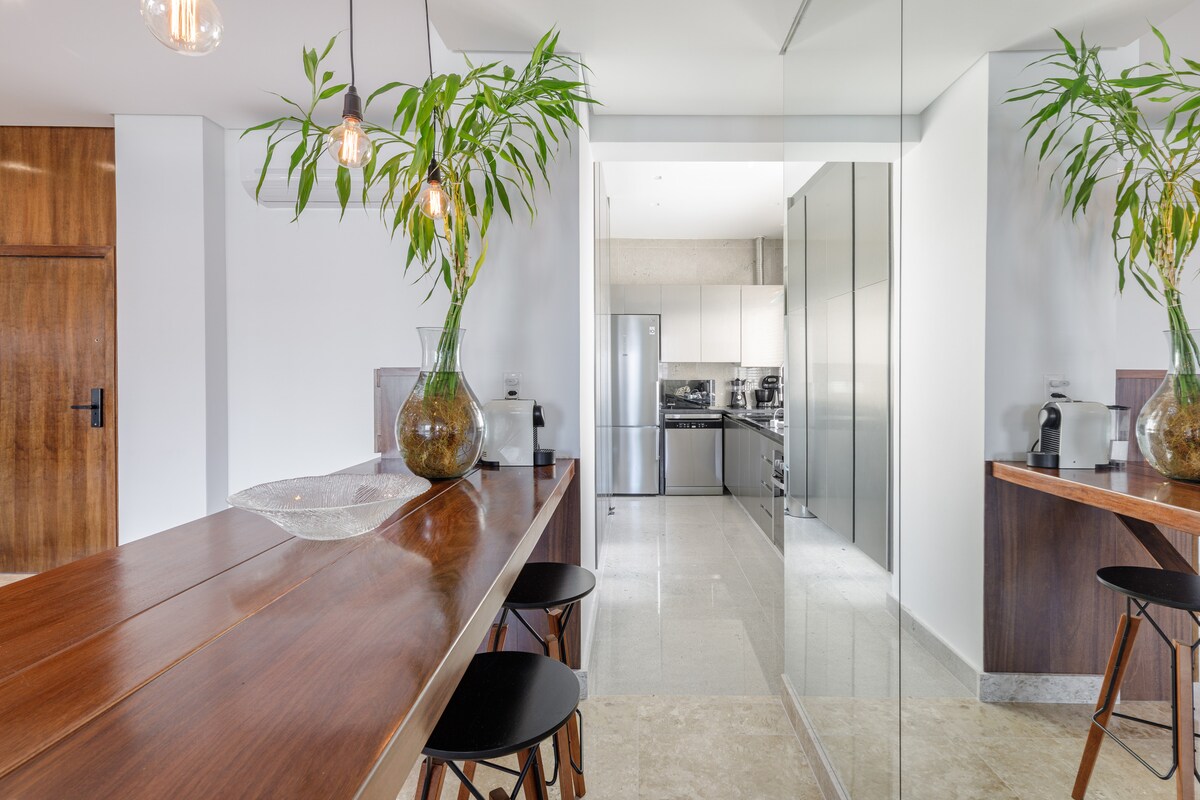 A modern kitchen area is complemented by a stylish wooden dining table with a glass centerpiece. Sleek cabinetry and stainless steel appliances are visible in the background. Natural light reflects off mirrored surfaces, creating a bright and inviting space.