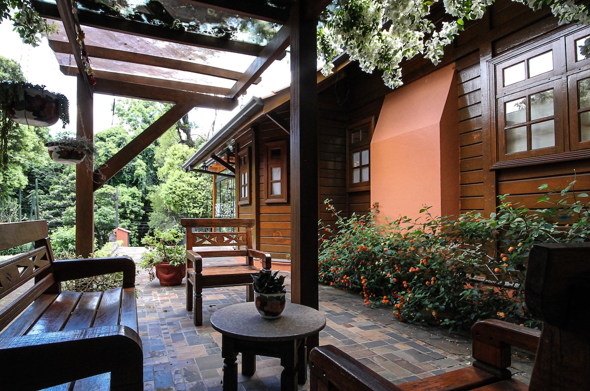 A wooden terrace is presented with natural stone flooring, featuring a round table surrounded by wooden chairs. Lush greenery and flowering plants are visible in the background, enhancing the serene atmosphere. The charming wooden structure is partially shaded by a lattice cover adorned with climbing plants.