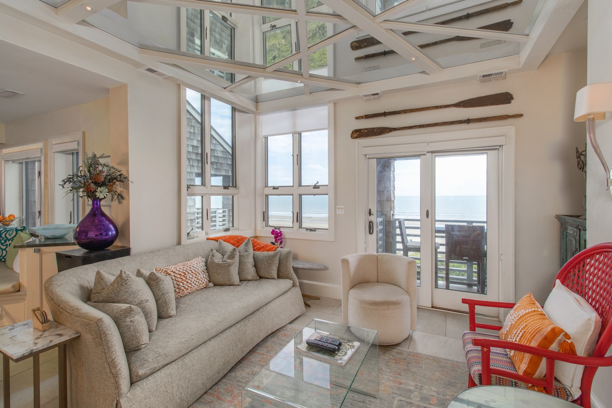 A comfortable living area features a spacious beige sofa adorned with vibrant throw pillows. A glass coffee table sits in front of the seating, while a red chair adds a pop of color. Natural light floods the room, highlighting coastal views beyond large windows.