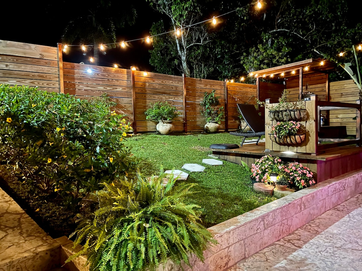 A serene garden area is highlighted by neatly arranged green grass and a wooden deck. Soft lighting from string lights casts a warm glow. Planters with foliage and flowers add color, while a seating area invites relaxation amidst the tranquil environment.