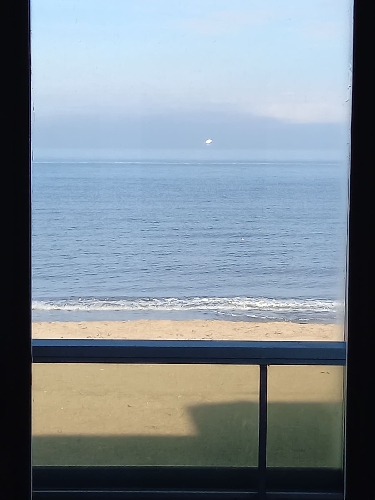A serene view of the ocean is framed by a closed loggia window. Gentle waves meet the sandy beach, while a distant horizon blends with the sky. The soft colors of the sea and sand are visible, creating a sense of tranquility.