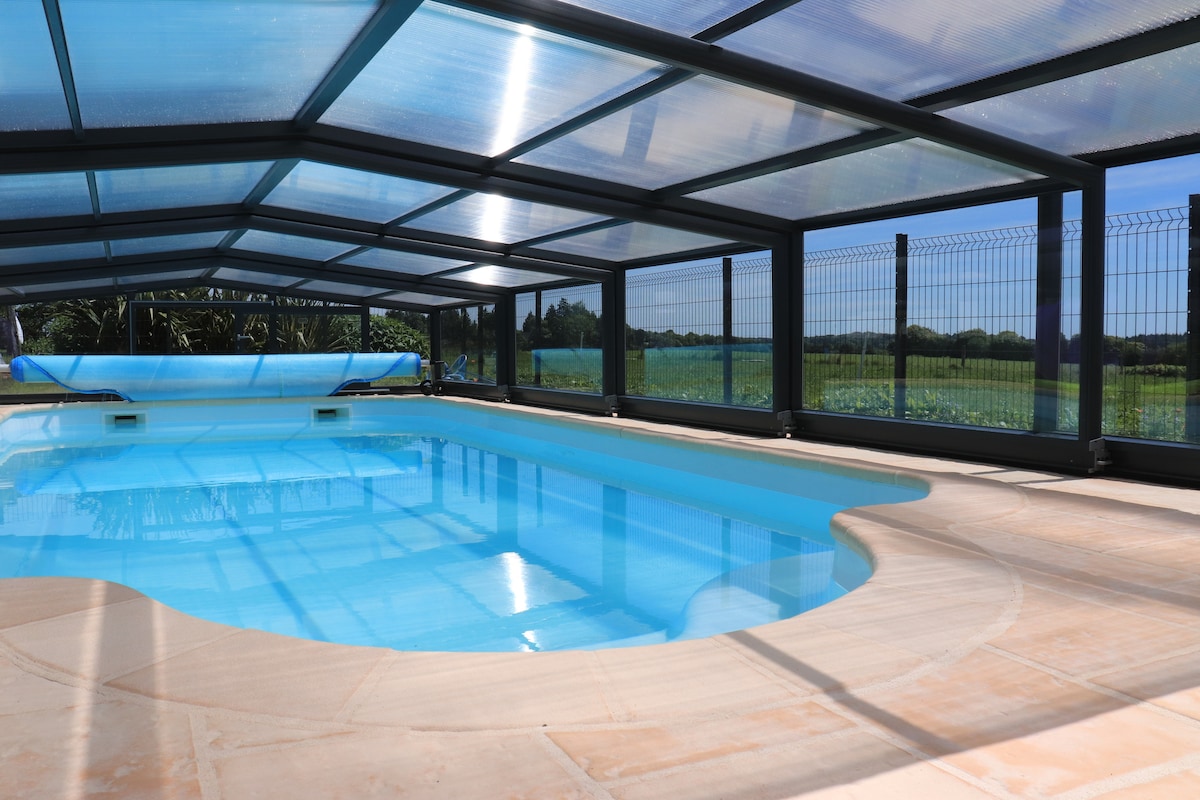 The indoor heated swimming pool is enclosed by glass panels, allowing natural light to illuminate the clear water. Surrounding views of greenery are visible through the transparent walls, creating a serene atmosphere. The pool area features a smooth, tiled surface and is designed for shared use.