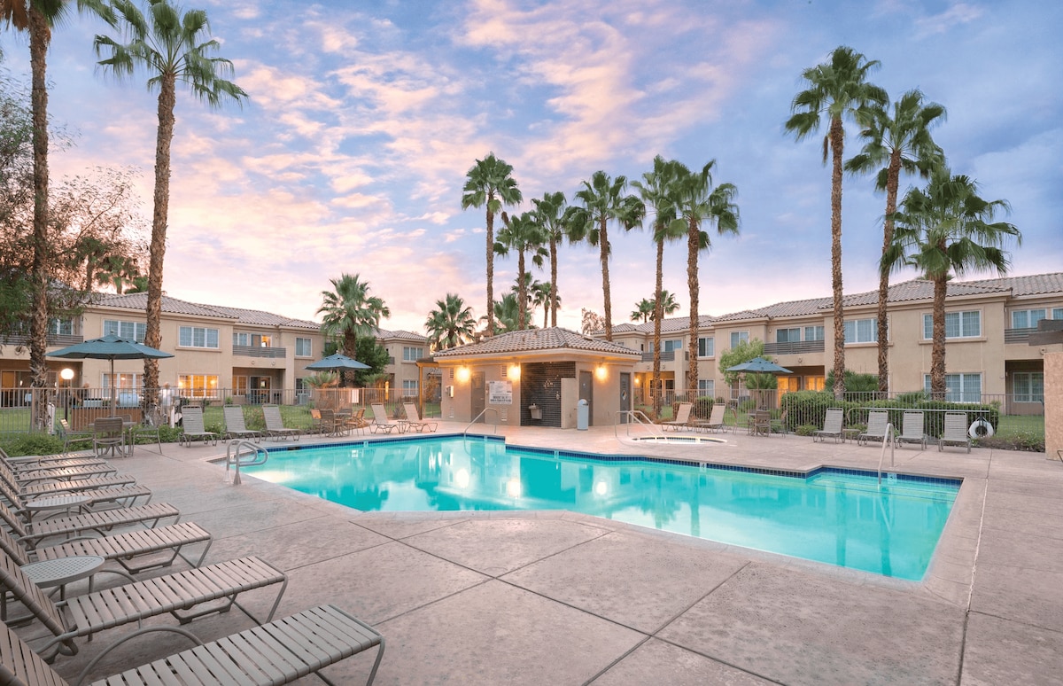 The outdoor pool area features a sparkling swimming pool surrounded by lounge chairs. Tall palm trees provide shade, and a covered seating area with picnic tables is available. The colorful sky at sunset enhances the inviting atmosphere of the space.