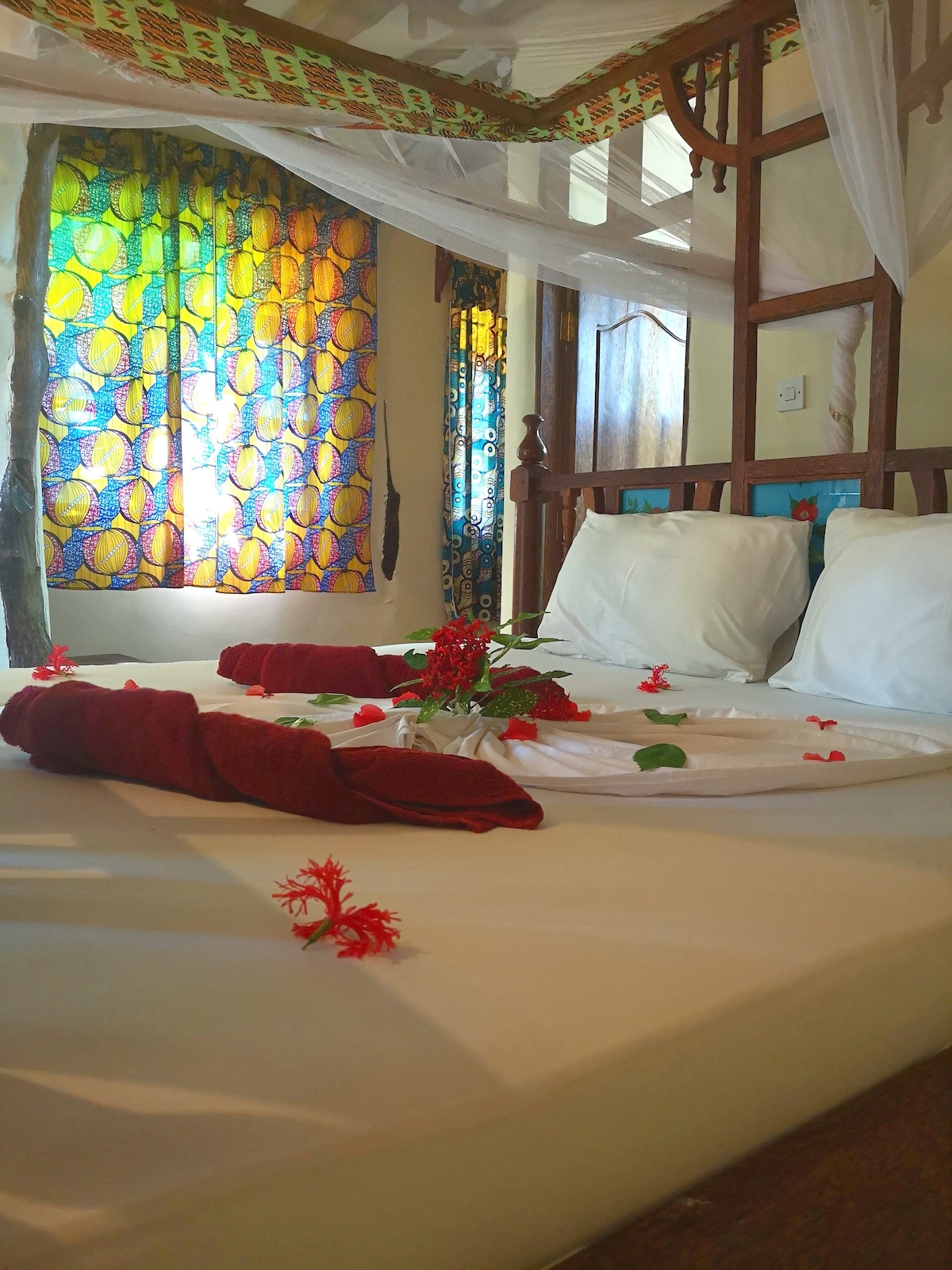 A single bed is adorned with white linens and decorated with red flowers. A mosquito net hangs above, while colorful patterned curtains filter natural light, adding vibrancy to the room. Simple wooden accents complement the serene ambiance, enhancing the inviting atmosphere.