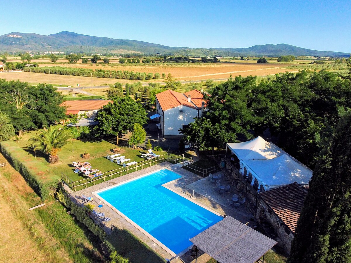 An aerial view captures the inviting pool surrounded by a spacious patio area with loungers. Lush greenery and trees frame the property, while nearby structures are visible, including a large gazebo for gatherings. Rolling hills and open fields stretch in the background.