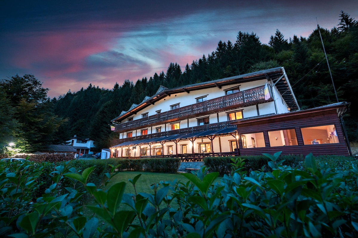 A multi-story lodge is positioned among lush greenery, featuring multiple balconies adorned with wooden railings. The evening sky presents a soft gradient of pink and blue hues. Warm lighting from the lower levels adds a welcoming glow, highlighting the serene natural surroundings.