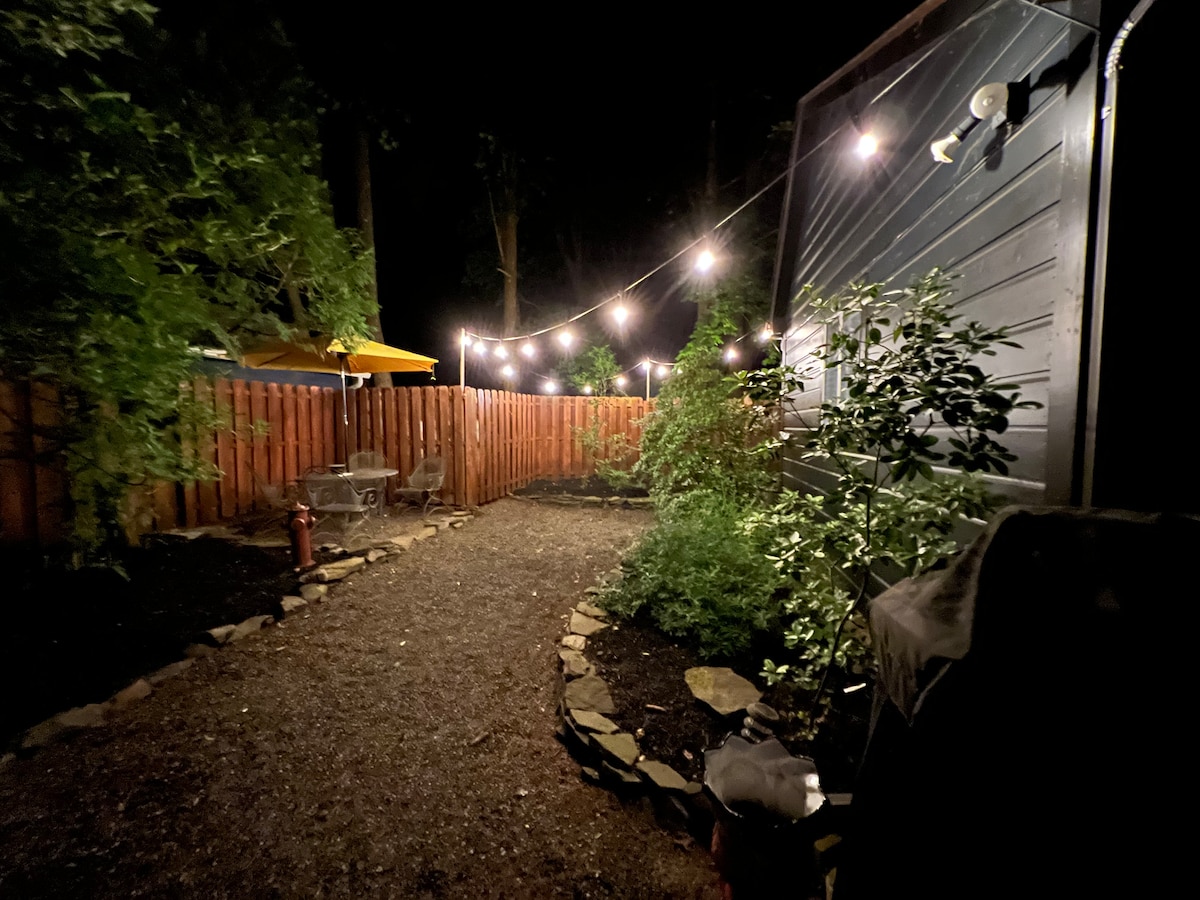 A gravel pathway winds through a lush yard, bordered by a privacy fence adorned with string lights. The garden features a fire pit area and a small table for outdoor seating, surrounded by greenery and illuminated by a warm glow from the lights.