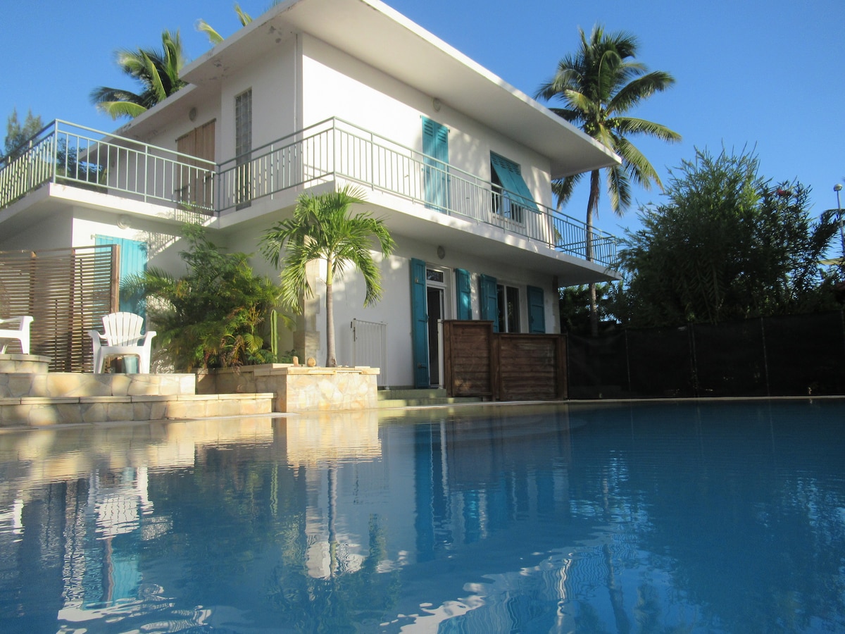 The villa features a two-story structure with turquoise shutters, surrounded by lush greenery. A spacious terrace overlooks a large pool, reflecting the clear blue sky. Sun loungers are positioned near the water's edge, creating a serene ambiance for relaxation.