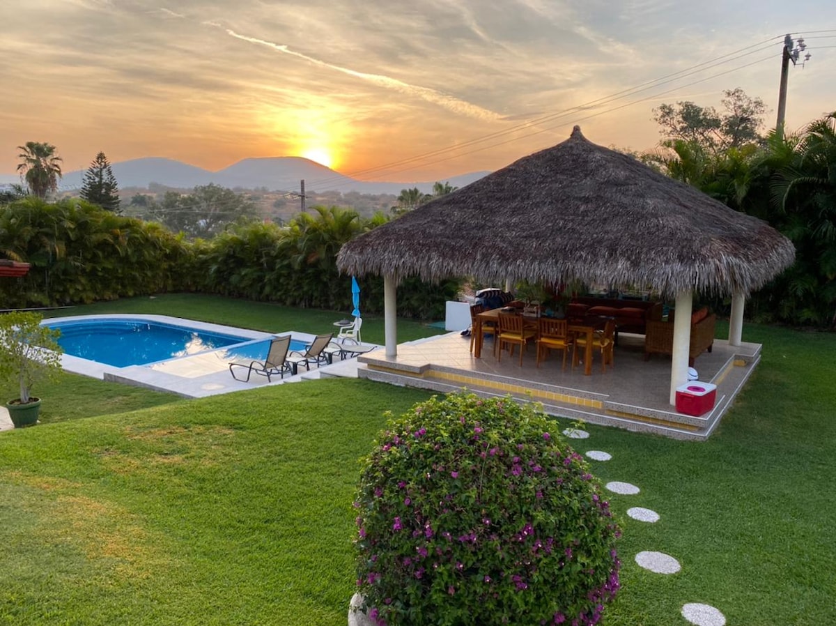 A lush green garden features a private pool surrounded by sun loungers. A palapa with a thatched roof offers shaded seating and dining space. A vibrant flower bush adds a pop of color, while a scenic sunset casts soft hues over the landscape.