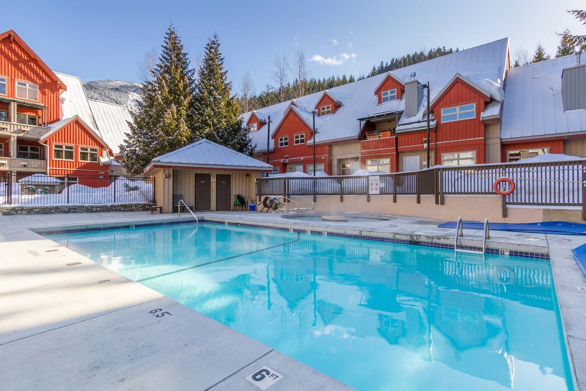 A heated outdoor pool is shown, surrounded by a wooden deck and lounge areas. Snow-covered trees stand in the background, alongside the multi-story red lodge buildings. The clear blue water reflects the bright sky, inviting relaxation year-round.