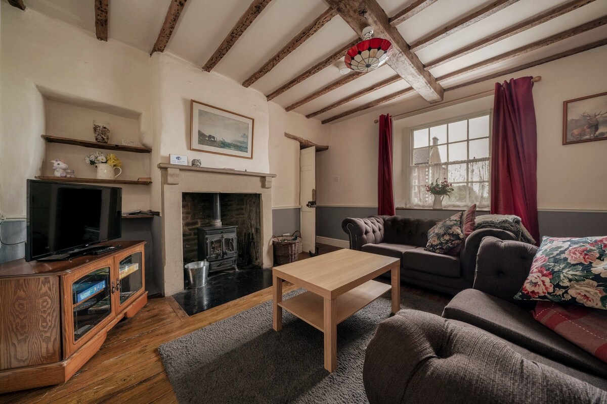 The lounge features wooden beams and a log burning stove, creating a rustic atmosphere. A coffee table sits at the center, surrounded by comfortable seating. Natural light is provided by a large window adorned with red curtains, and a television is positioned beside a wooden cabinet.