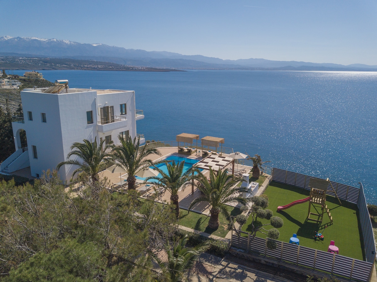 An aerial view illustrates the villa set against a stunning seascape, featuring a spacious pool surrounded by sunbeds. Lush palm trees and a play area are visible in the meticulously landscaped yard, with the distant mountains framing the coastal horizon.