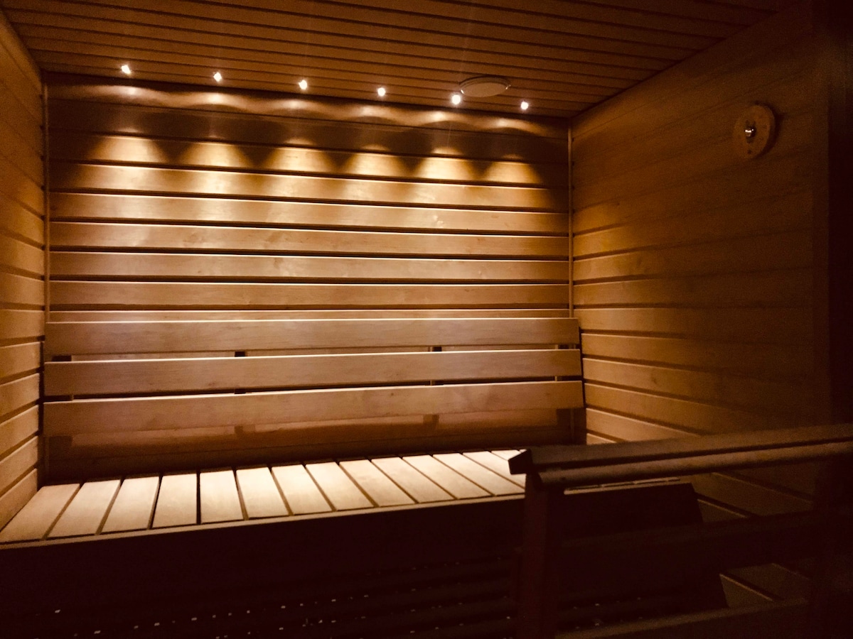 A wooden sauna interior is depicted, featuring horizontal wooden planks on the walls and benches. Soft, ambient lighting is installed along the upper edge, creating a warm glow. The space is well-structured, emphasizing a tranquil and relaxing atmosphere.