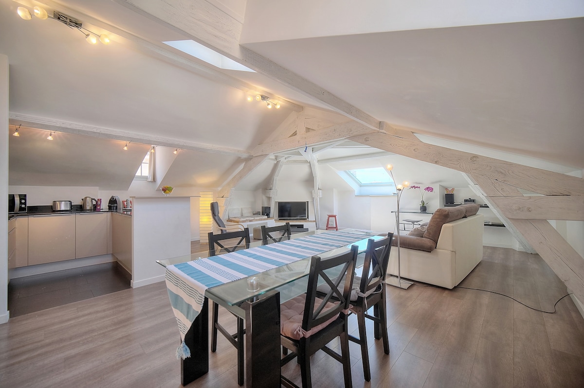 The open-plan space features a dining table with dark wooden chairs, illuminated by ceiling lights. A comfortable sofa is positioned nearby, with the sloped ceiling revealing wooden beams. A fully equipped kitchen is visible in the background, adding to the modern and functional appeal.