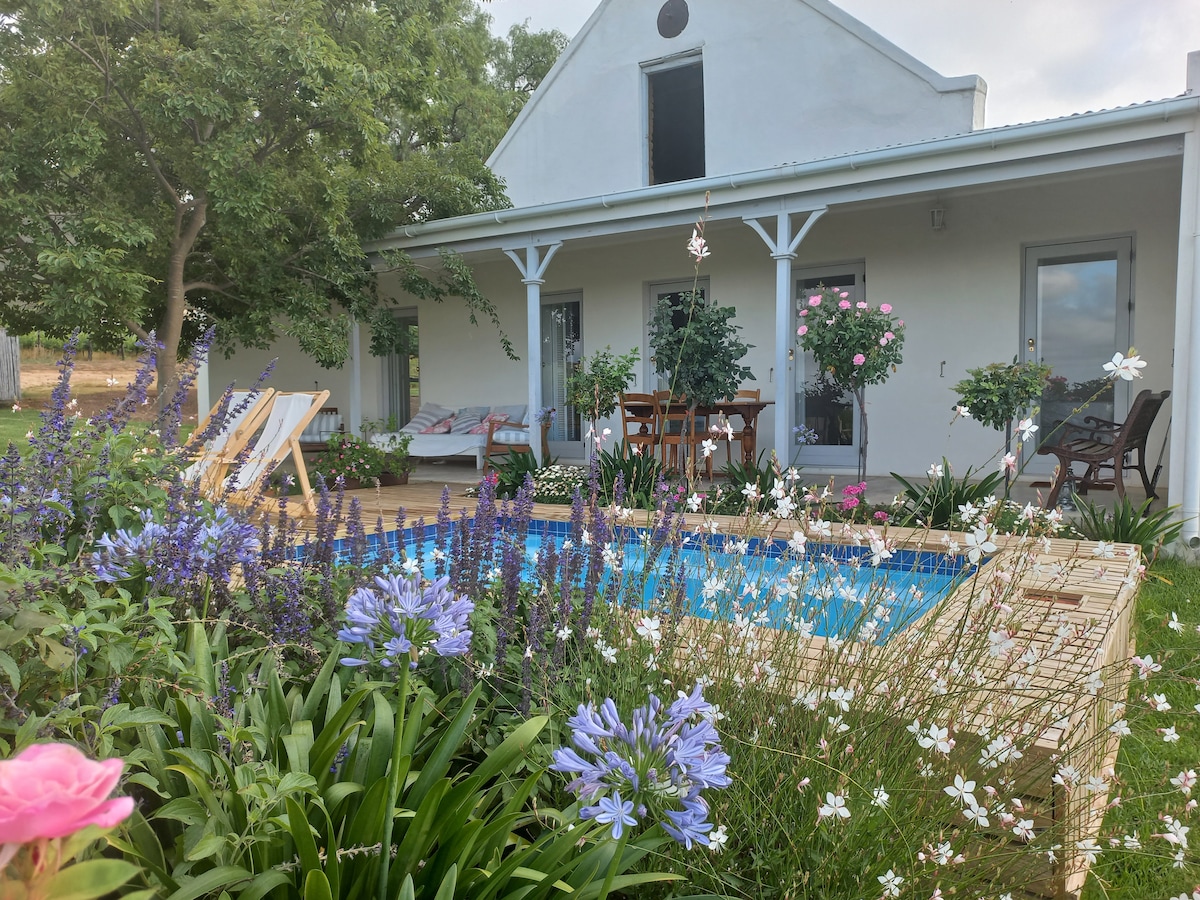 A charming outdoor area features a private pool surrounded by lush greenery and colorful flowers. The cottage’s patio includes a small dining table and comfortable seating. Planter boxes with hanging flowers add a floral touch, while sun loungers invite relaxation in the serene setting.