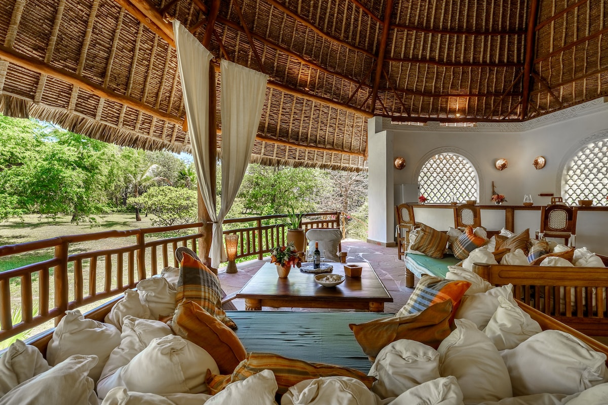 A spacious seating area features plush cushions on wooden benches, designed for comfort. Light fabric drapes enhance the relaxed atmosphere, while large windows frame views of the lush garden outside. A low wooden table occupies the center, complemented by decorative items.