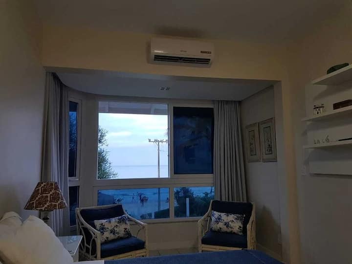 Quarto 01 (Suíte voltada para a praia): vista do painel com TV de LED, ar condicionado,  poltronas e amplas janelas com vista para o mar
