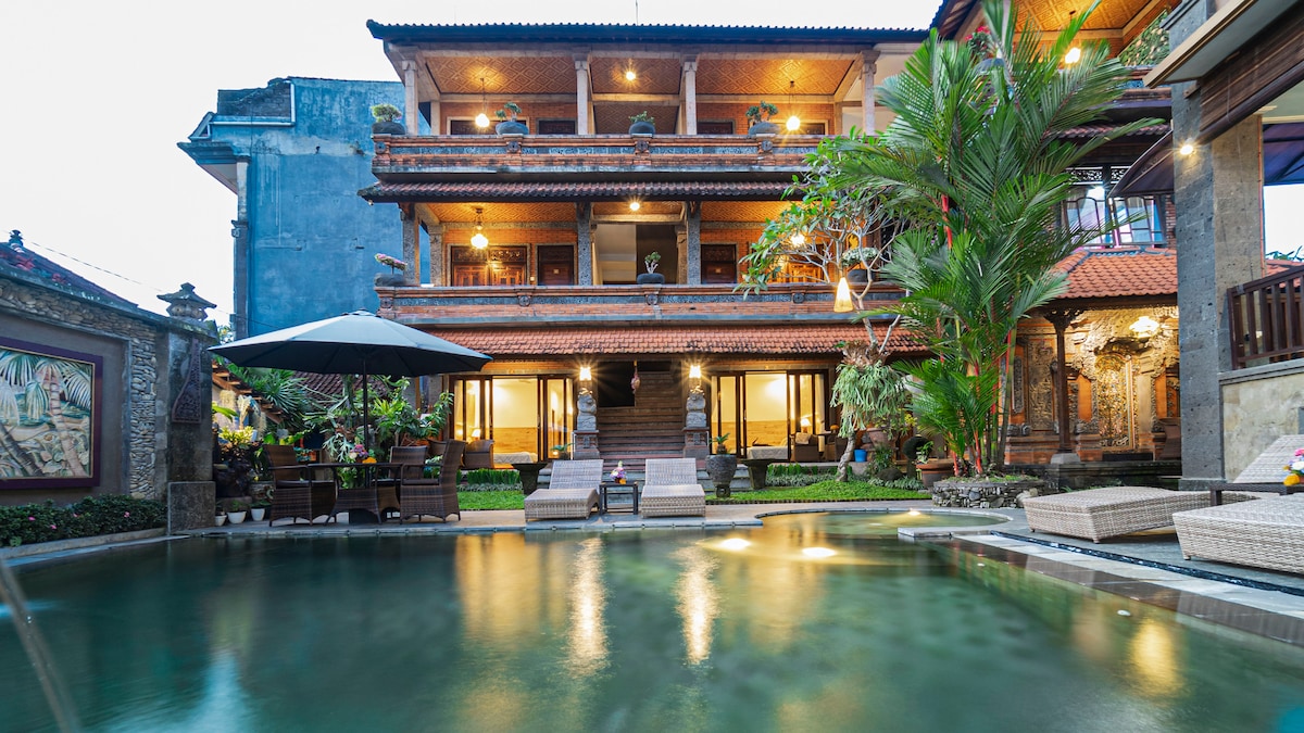 The exterior of Kun - Kun Guest House 3 showcases a multilevel structure surrounded by lush greenery. A swimming pool is visible in the foreground, reflecting warm lights from the building. Outdoor seating areas are arranged under umbrellas, enhancing the inviting atmosphere of the property.