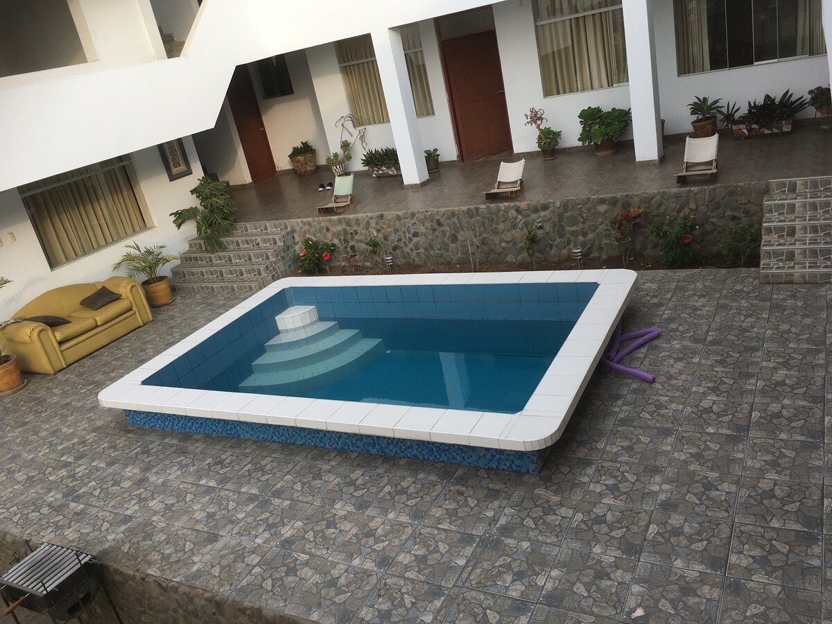 A clean, rectangular swimming pool is displayed in the courtyard, surrounded by a tiled terrace. The pool features steps leading into the water and is framed by plants and lounge chairs. The adjacent area includes a comfortable seating arrangement and light-colored walls.
