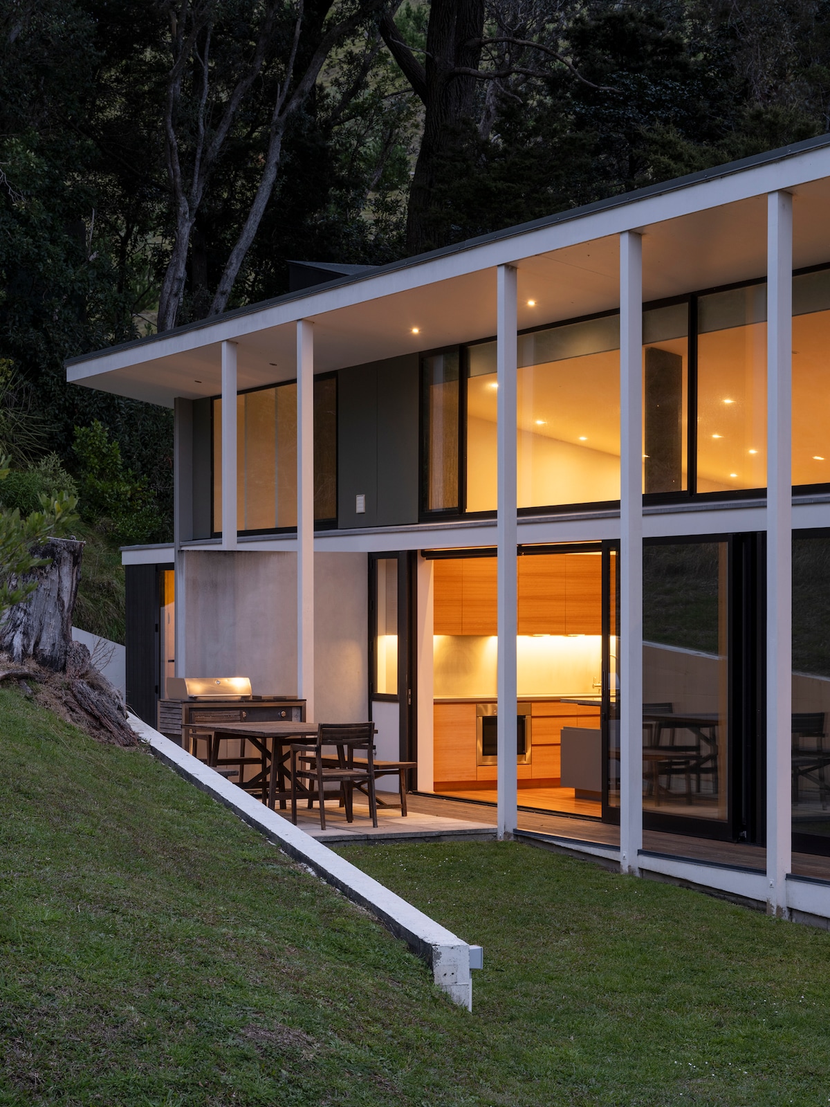 The contemporary structure features large glass walls, allowing natural light to illuminate the interior spaces. A patio area with wooden furniture is visible, set against a backdrop of lush greenery and gentle slopes. Soft lighting highlights the kitchen area within sight of the exterior.