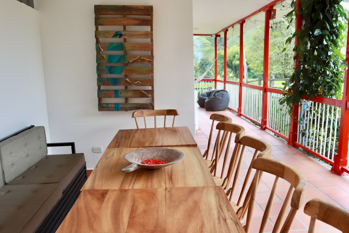 A dining area is presented with a rustic wooden table surrounded by several chairs. A decorative bowl with red accents is centered on the table. A vibrant piece of wall art featuring a bird is visible, complemented by greenery along the balcony railing.