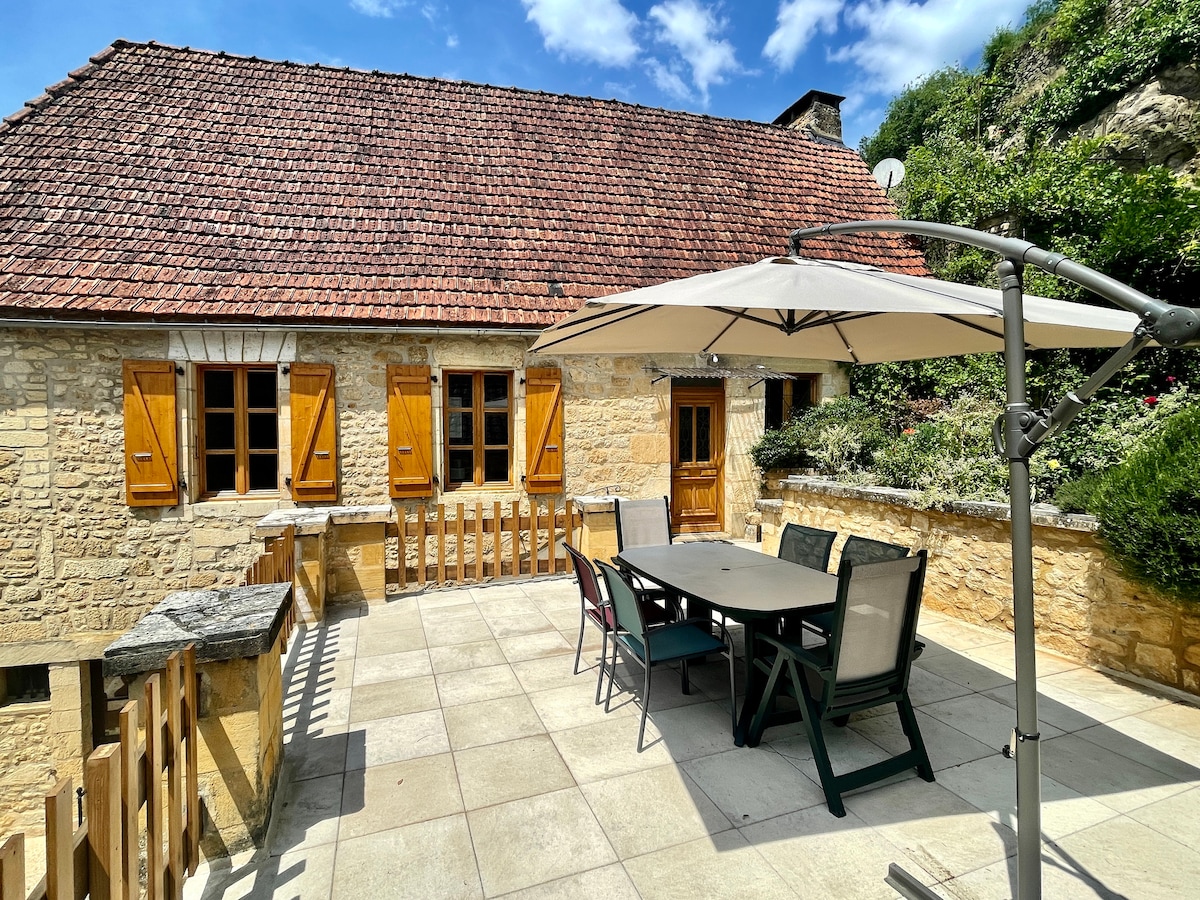 An outdoor terrace features a large dining table surrounded by chairs, shaded by a sun umbrella. Natural stone walls and window shutters in warm hues complement the tranquil setting, which is bordered by lush greenery and a neatly maintained garden area.